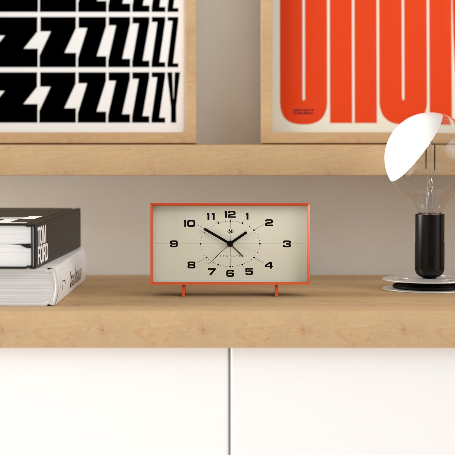 Eine rechteckige Uhr auf einem Holzregal mit Büchern, einer Lampe und bunt gerahmten Postern an der Wand.