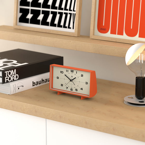 Orangefarbene Retro-Uhr auf einem Holzregal, daneben Bücher und eine kleine Lampe mit runder Glühbirne.