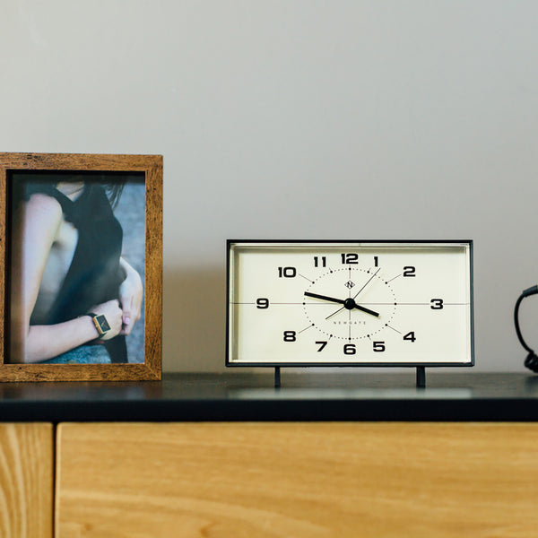 Ein gerahmtes Foto und eine rechteckige Analoguhr stehen auf einer Holzkommode an einer schlichten Wand.