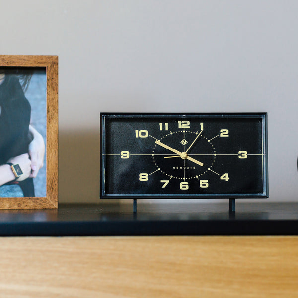 Eine schwarze Vintage-Uhr, die 12:55 Uhr anzeigt, steht auf einem Holzregal neben einem gerahmten Foto.