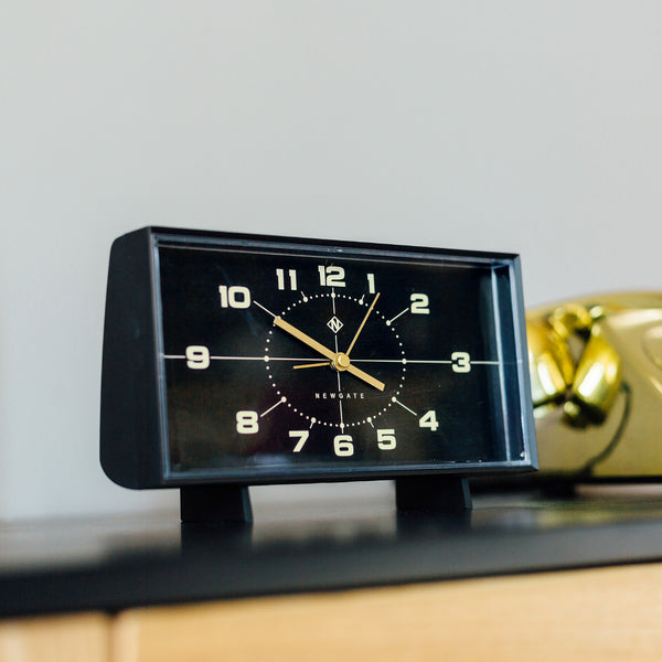 Schwarze Analoguhr im Retrostil, die 10:10 anzeigt und auf einer dunklen Fläche neben einem goldenen Objekt steht.