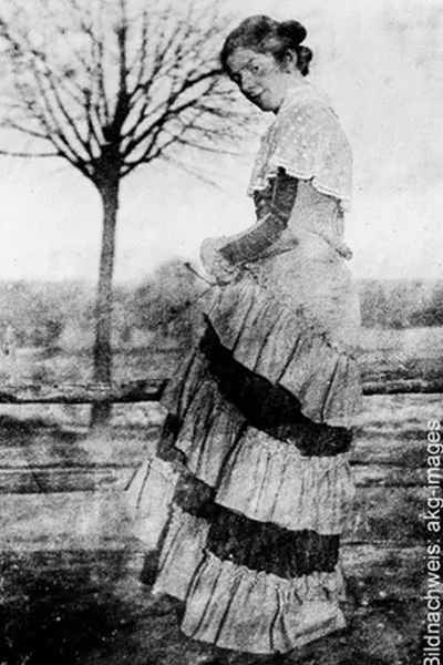Eine Frau in einem langen, mehrlagigen Kleid steht im Freien an einem Holzzaun, und im Hintergrund ist ein blattloser Baum zu sehen.