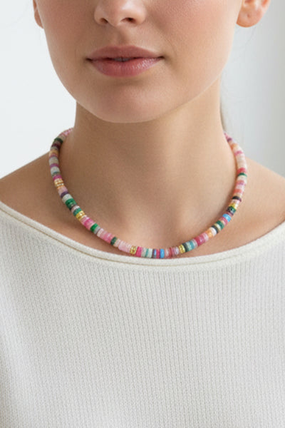 Eine Frau, die ein geripptes weißes Oberteil und eine bunte Perlenkette trägt, wird von den Schultern aufwärts vor einem schlichten Hintergrund gezeigt.