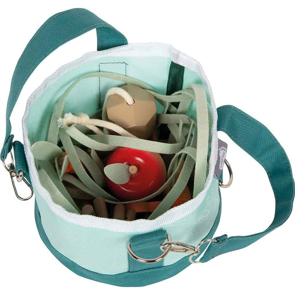 Eine grüne Stofftasche, gefüllt mit Spielzeuggemüse aus Holz, darunter ein roter Apfel und grüne Blätter, von oben betrachtet.