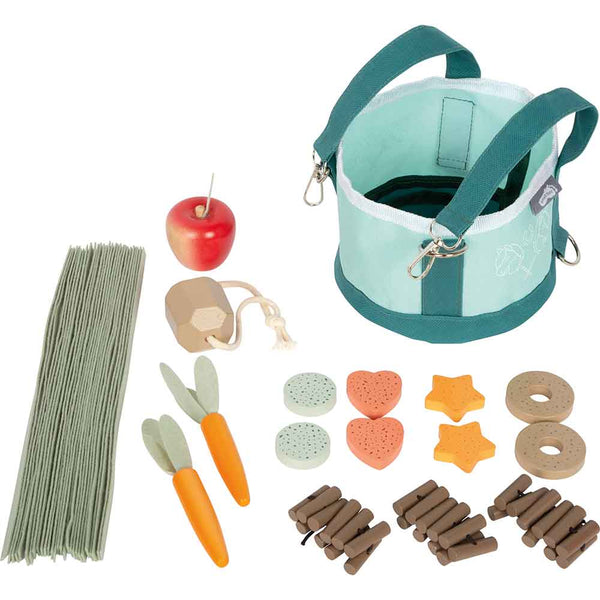 Ein Spielzeugset mit einem grünen Stoffbeutel einem Spielzeugapfel, einer Karotte, Heusträhnen, Holzscheiten und verschiedenen Spielzeugleckereien, die auf einer weißen Fläche angeordnet sind.