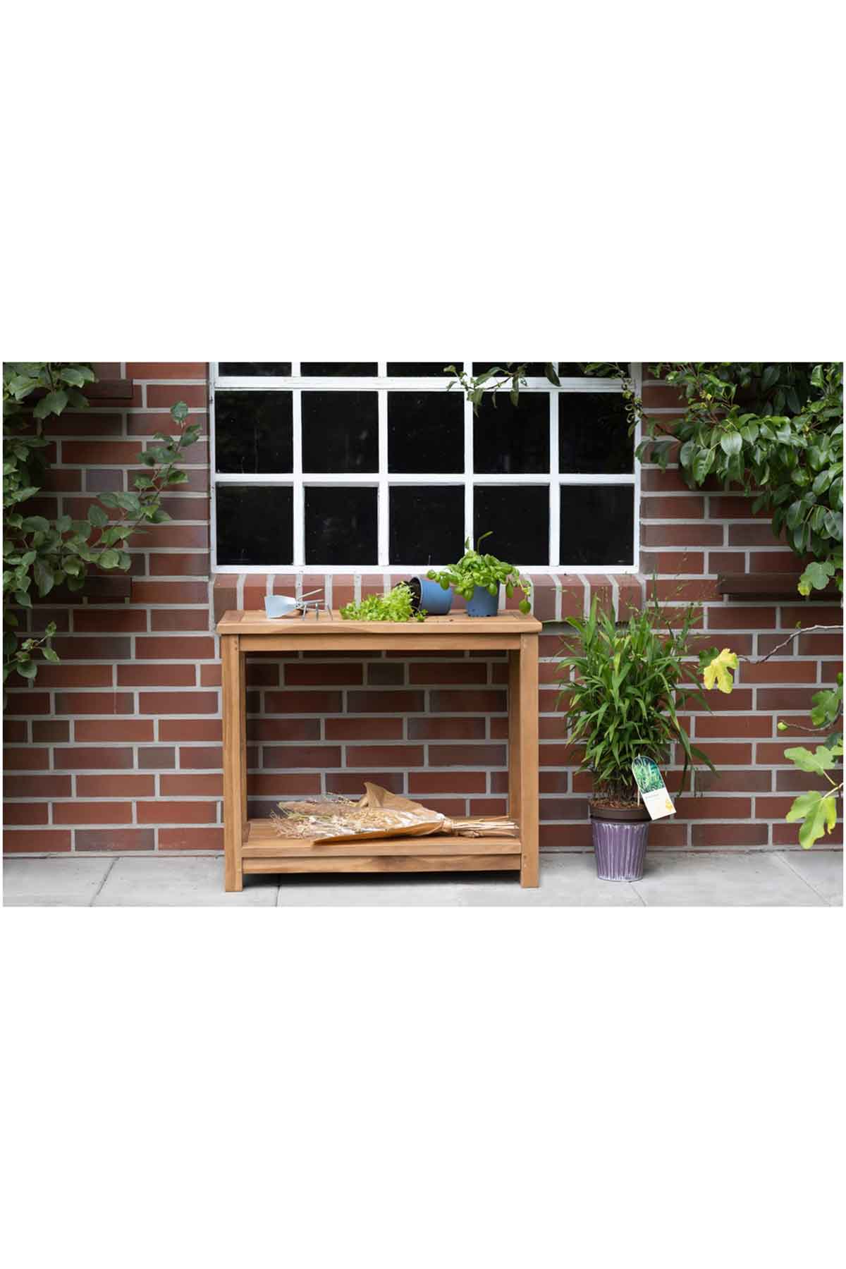 Vor einer Backsteinmauer und einem Fenster steht ein Holztisch mit Pflanzen, Kräutern und Gartengeräten, rechts daneben eine Topfpflanze.