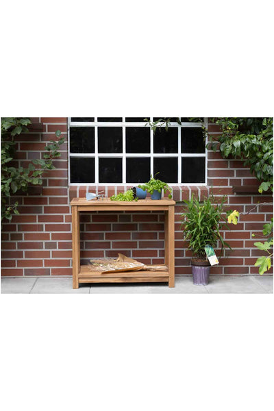 Vor einer Backsteinmauer und einem Fenster steht ein Holztisch mit Pflanzen, Kräutern und Gartengeräten, rechts daneben eine Topfpflanze.