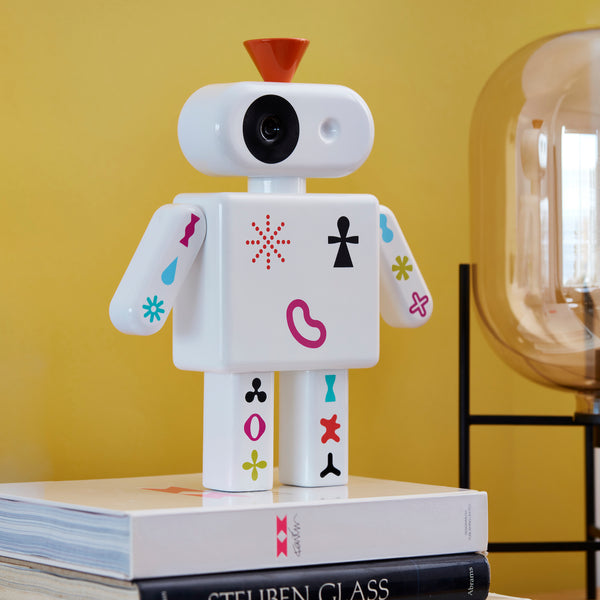 Ein Spielzeugroboter mit bunten Symbolen steht auf gestapelten Büchern neben einer Glaslampe vor einer gelben Wand.
