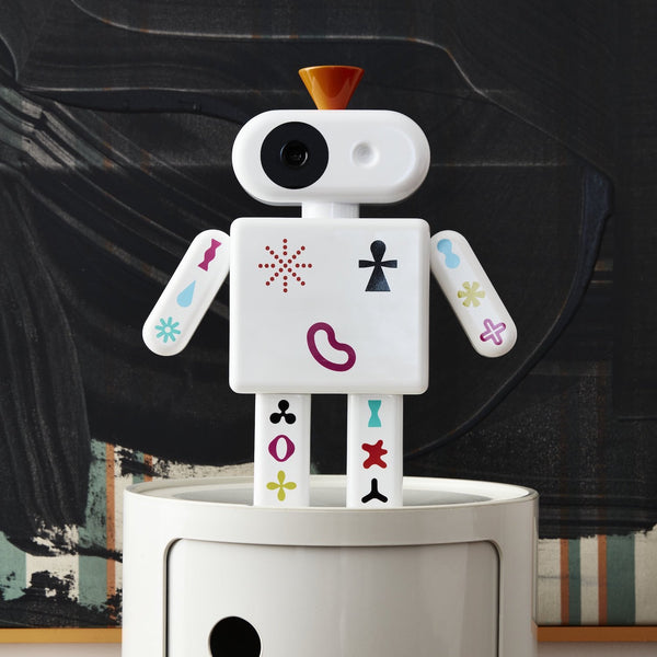 Ein Spielzeugroboter mit bunten Symbolen steht auf einer weißen Fläche vor einem dunkel gemusterten Hintergrund.