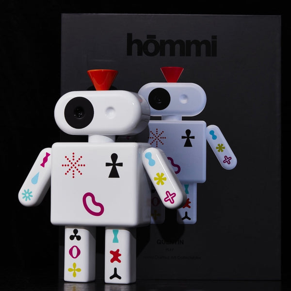 Ein weißer Spielzeugroboter mit bunten Symbolen steht vor einer schwarzen Schachtel mit der Aufschrift hömmi.