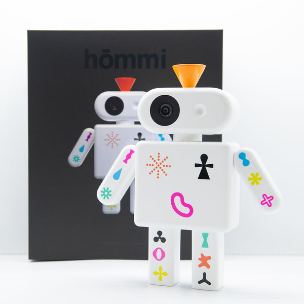 Ein weißer Spielzeugroboter mit bunten Formen steht vor einer schwarzen Schachtel, auf der sein passendes Bild und das Wort hōmmi abgebildet sind.
