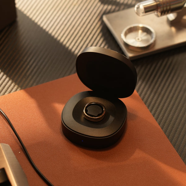 Ein intelligenter Ring liegt in einer offenen Ladeschale auf einem braunen Schreibtisch mit einem Kabel in der Nähe.