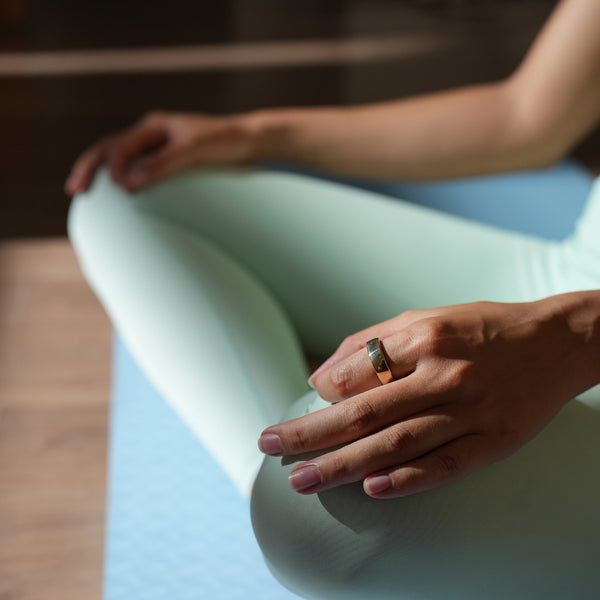 Person, die im Schneidersitz auf einer Yogamatte sitzt, hellblaue Leggings trägt und einen Ring an der Hand hat.