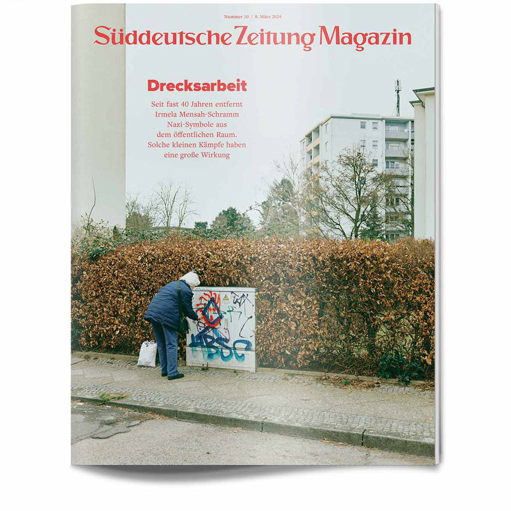 Eine Person in einer blauen Jacke entfernt Graffiti von einem Stromkasten in der Nähe einer braunen Hecke in einem städtischen Gebiet.