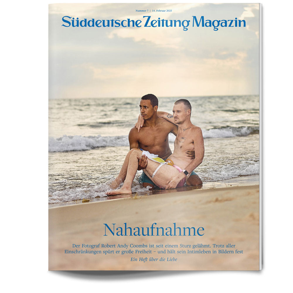 Zwei Männer sitzen zusammen am Strand, der eine auf dem Schoß des anderen, mit den Wellen im Hintergrund.