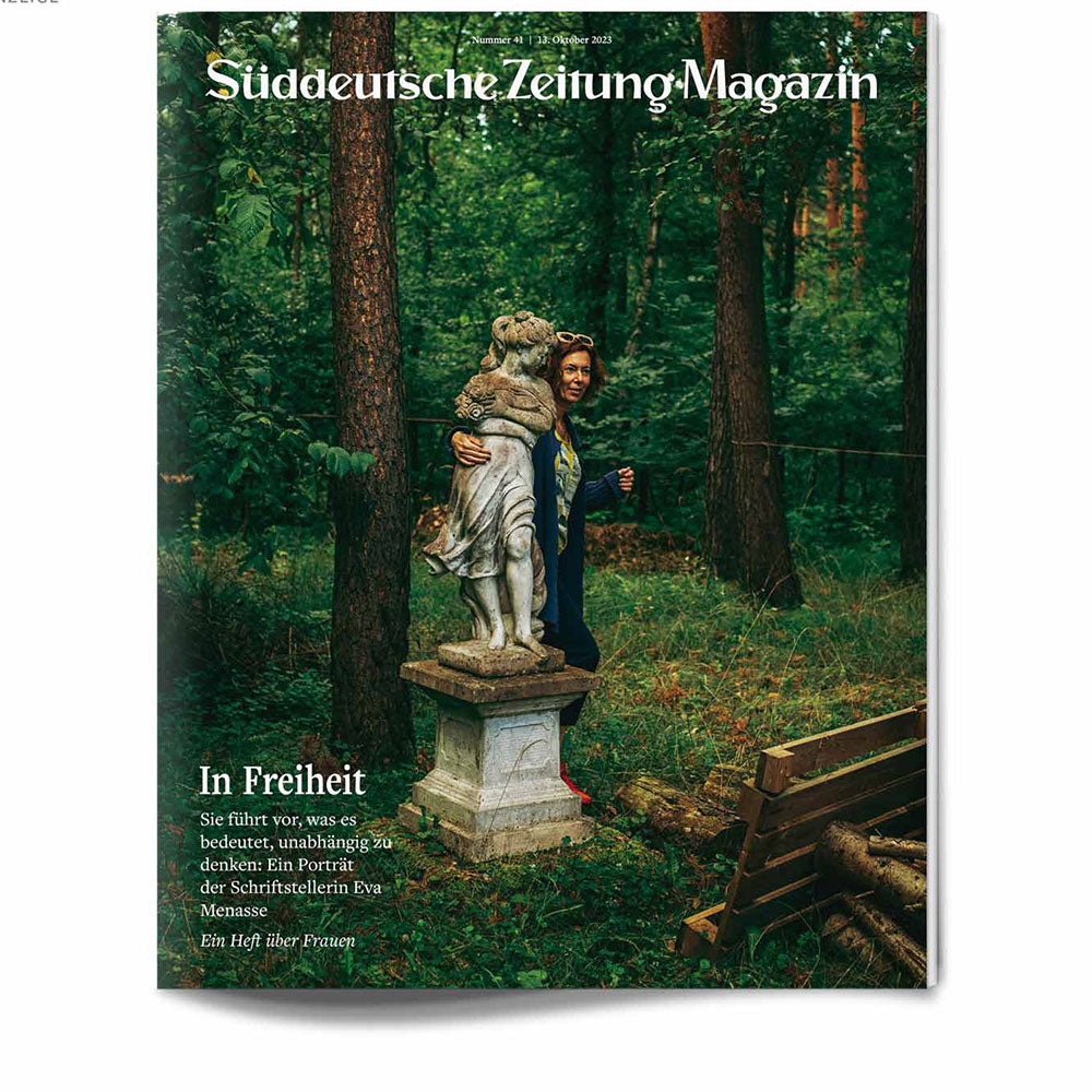 Eine Frau steht in einem Wald und stützt sich auf eine steinerne Bärenstatue; im Hintergrund sind Bänke und Bäume zu sehen.