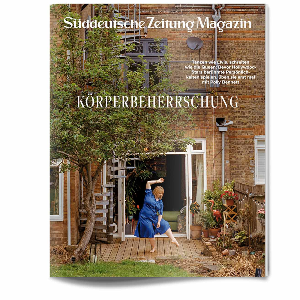 Eine Frau in Blau posiert auf einer Holzterrasse vor einem Backsteinhaus, umgeben von Grün.