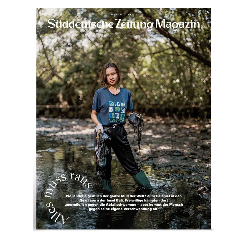 Eine Frau steht mit Stiefeln, Handschuhen und einem T-Shirt mit Recycling-Symbolen in einem schlammigen Fluss und hält Müll.