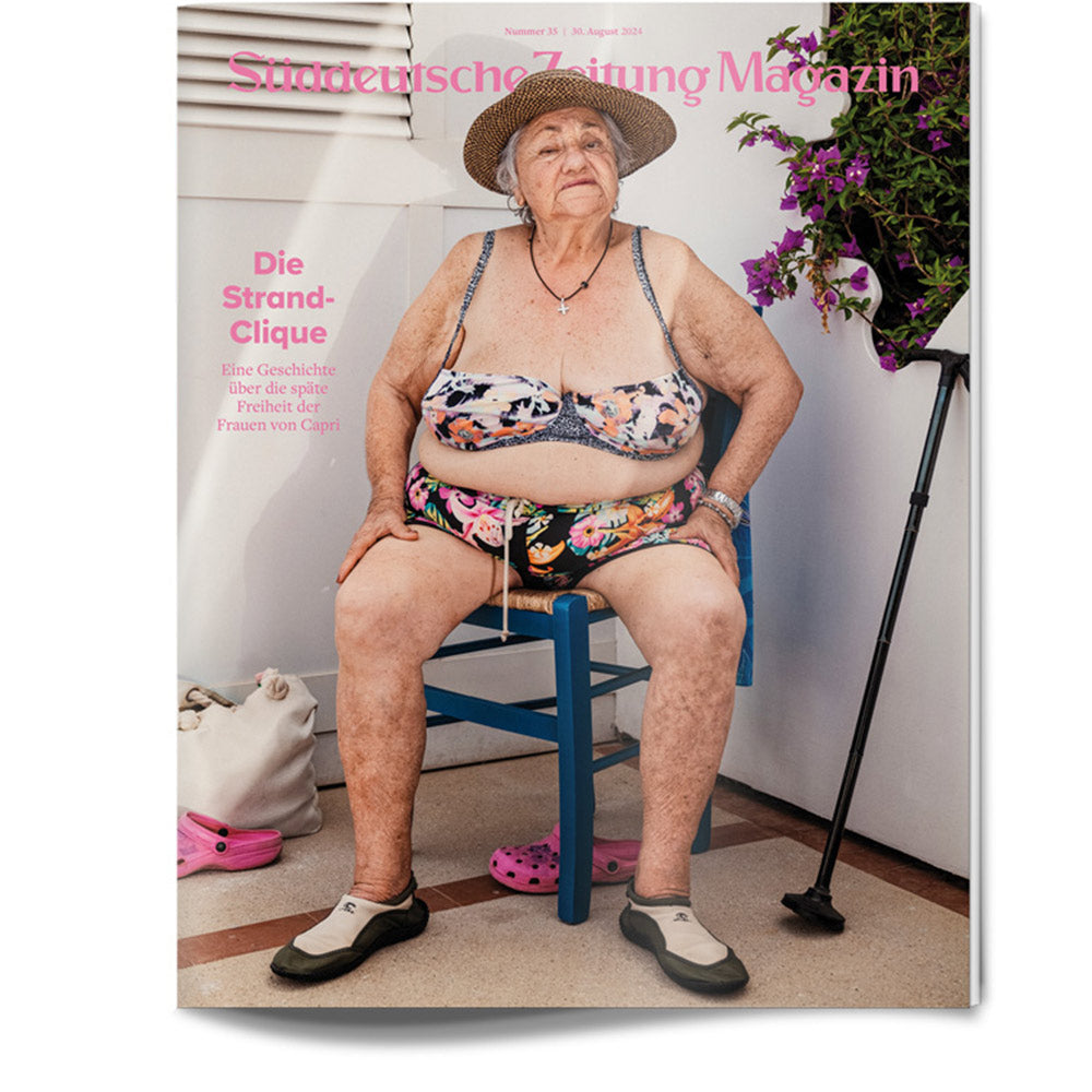 Ältere Frau im Bikini und mit Hut sitzt auf einem blauen Stuhl in einem Haus, in dessen Nähe Pflanzen und Schuhe stehen.