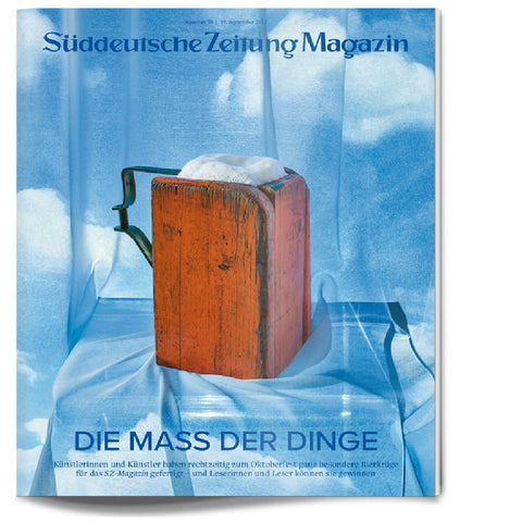 Ein mit Schaum gefüllter hölzerner Bierkrug steht auf einem drapierten blauen Tuch vor einem wolkenverhangenen Hintergrund.