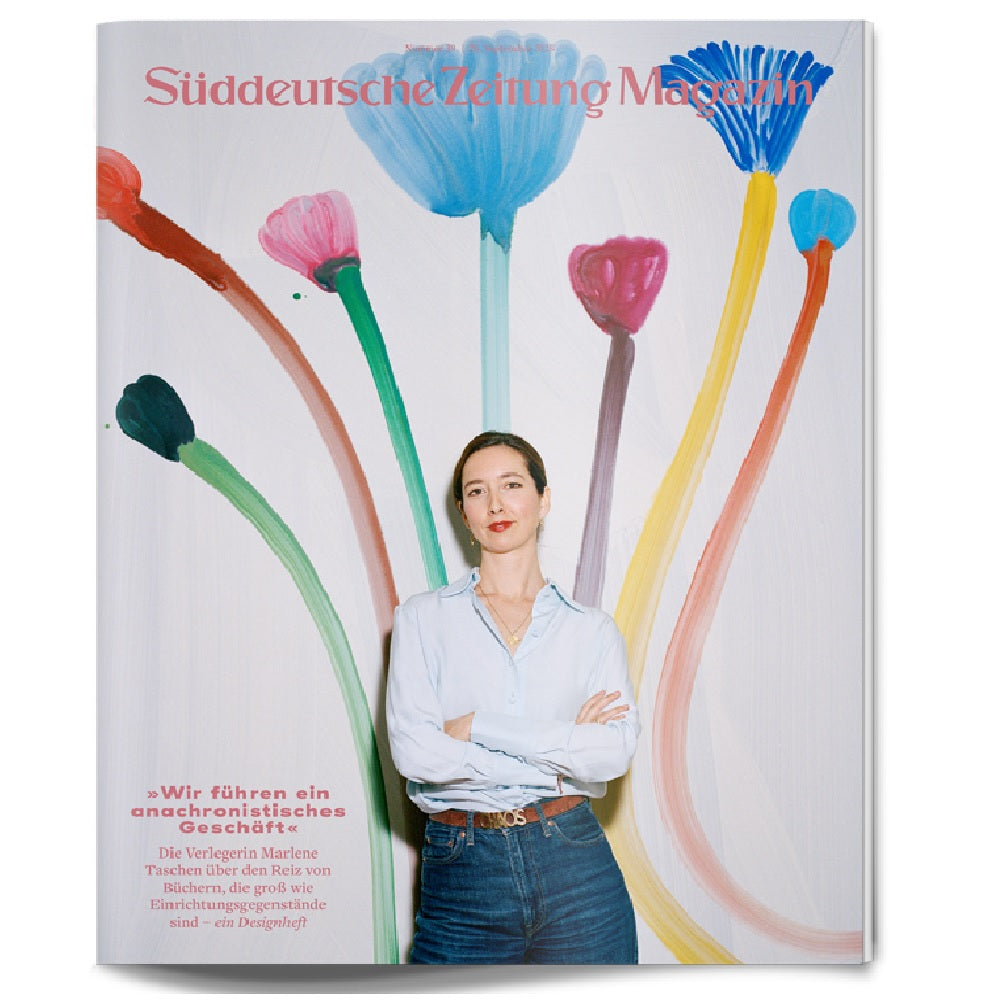 Frau in hellblauem Hemd steht vor einer Wand mit bunt bemalten Blumen auf dem Cover einer Zeitschrift.