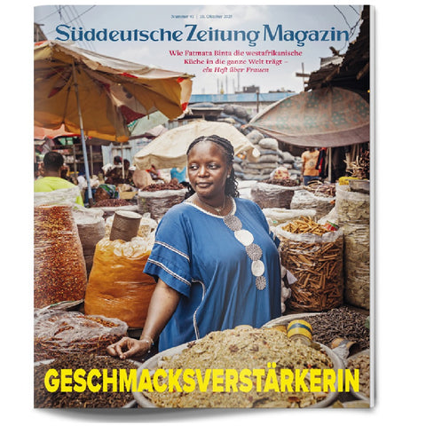 Eine Frau steht lächelnd auf einem belebten Markt im Freien, umgeben von Körben mit Lebensmitteln und Gewürzen.