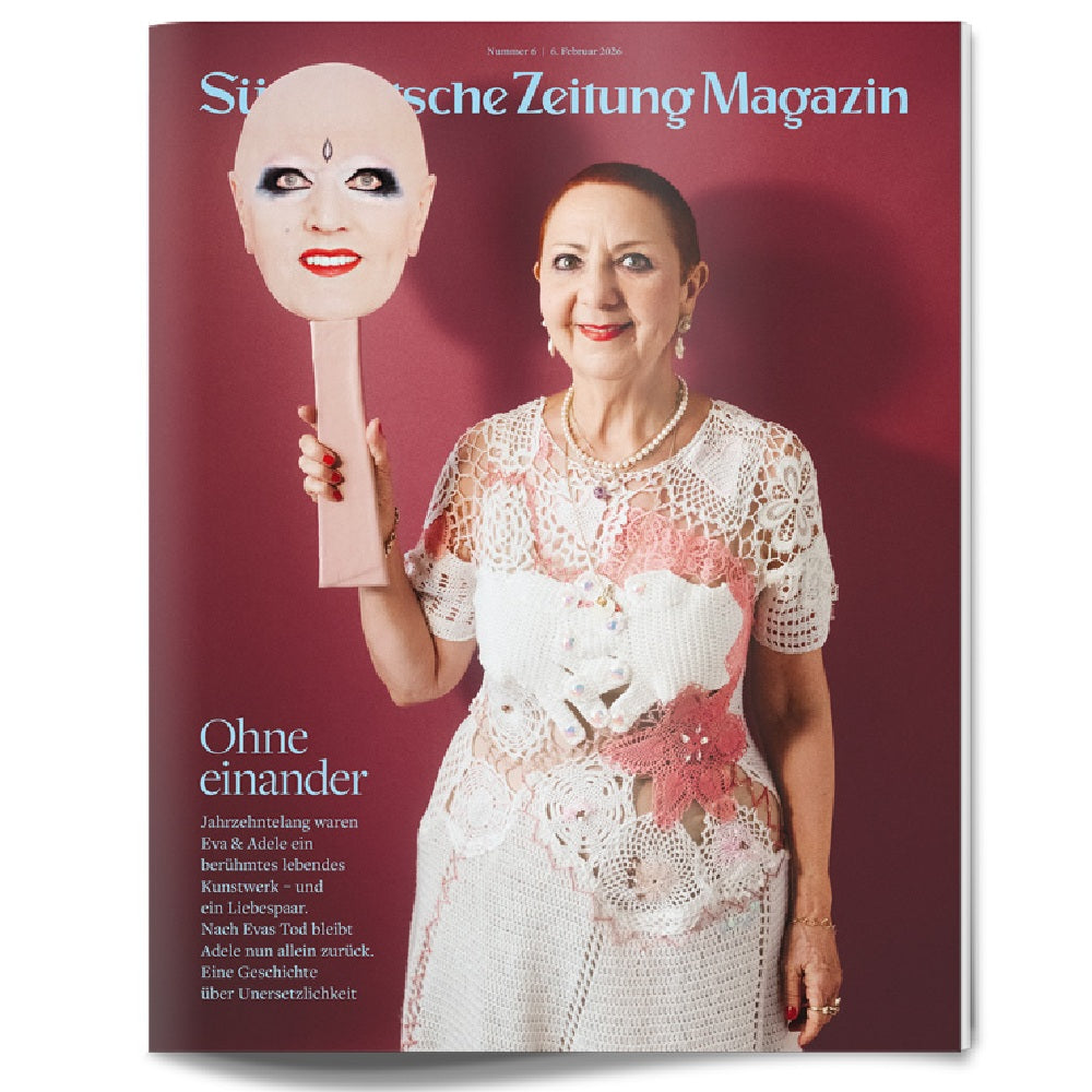 Eine Frau in einem Spitzenkleid hält eine große Maske mit einem gemalten, lächelnden Gesicht vor einem roten Hintergrund.