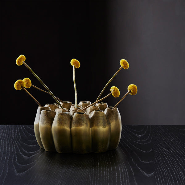 Eine skulpturale Vase mit gelben Billy-Ball-Blumen auf einem dunklen Holztisch vor einem schwarzen Hintergrund.