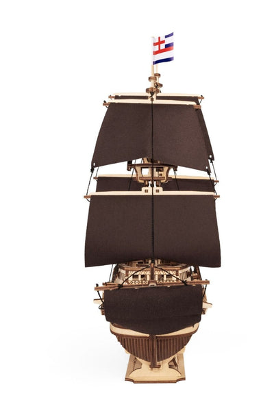 Ein Modell eines hölzernen Segelschiffs mit drei dunkelbraunen Segeln und einer Flagge mit einem roten Kreuz und blauen Streifen auf der Oberseite, von vorne gesehen vor einem weißen Hintergrund.