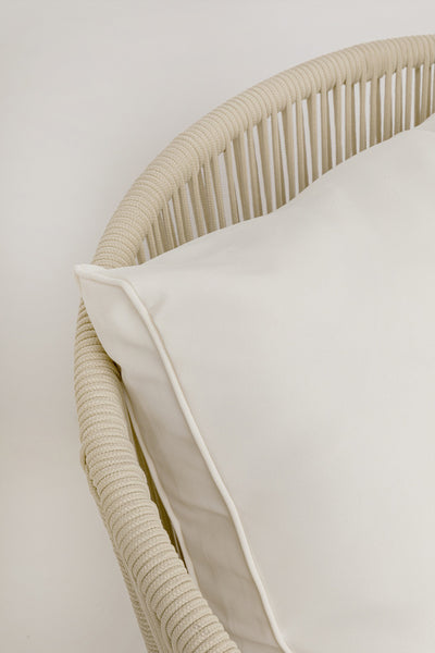 Nahaufnahme eines cremefarbenen Kissens auf einem geflochtenen, hellbeigen Stuhl mit seilartiger Textur vor einem unifarbenen Hintergrund.