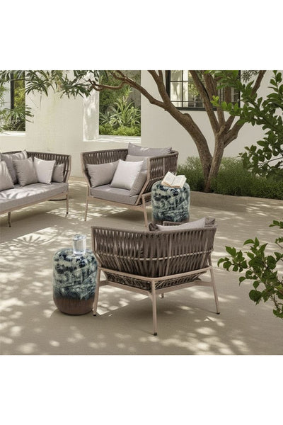 Terrasse im Freien mit Korbmöbeln, hellen Kissen, zwei Beistelltischen aus Keramik und üppigen grünen Pflanzen und Bäumen im Hintergrund.