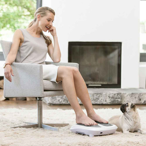Frau benutzt ein Fußmassagegerät in einem Wohnzimmer, während ein kleiner Hund sie anschaut.