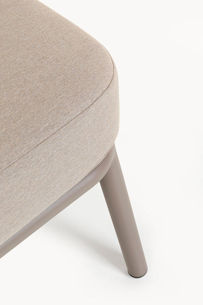 Nahaufnahme der Ecke eines beigefarbenen gepolsterten Stuhls mit einem abgerundeten Kissen und einem einzelnen angewinkelten, taupefarbenen Bein auf weißem Hintergrund.