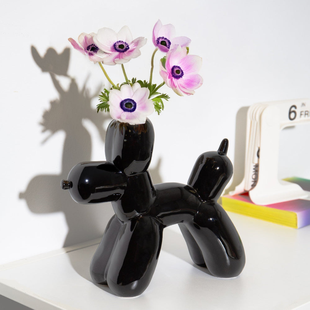Eine schwarze Vase in Form eines Ballonhundes hält rosa und weiße Blumen auf einer weißen Fläche neben einem blätterbaren Kalender und bunten Notizblöcken.