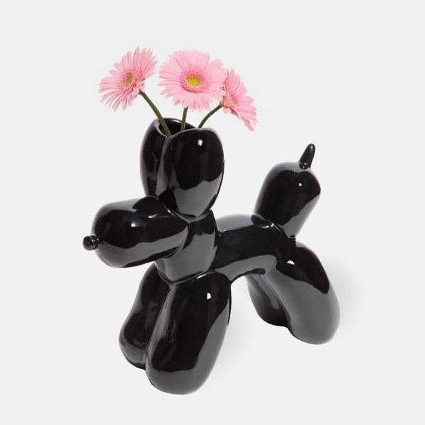 Eine schwarze Keramikvase in Form eines Ballonhundes hält zwei rosafarbene Blumen, die vor einem schlichten weißen Hintergrund stehen.