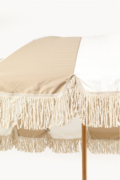 Ein beige-weiß gestreifter Sonnenschirm mit einer Holzstange und einer Fransenborte am Rand.