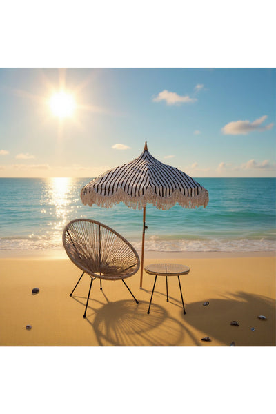 Ein runder Strohsessel, ein kleiner Tisch und ein gestreifter Sonnenschirm stehen an einem Sandstrand mit Muscheln und blicken auf ein ruhiges Meer unter einem sonnigen Himmel.