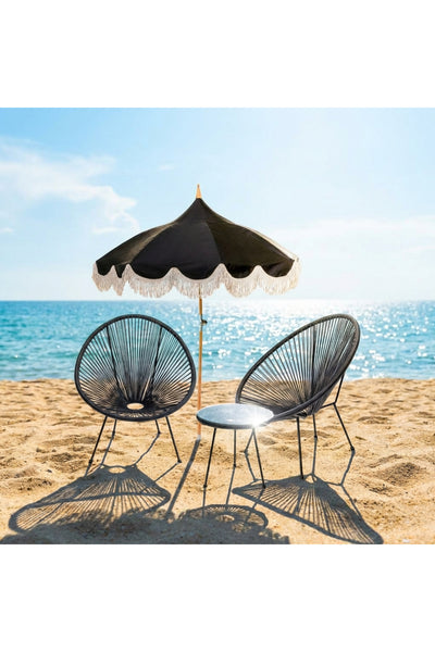Zwei schwarze Stühle und ein passender runder Tisch stehen unter einem schwarzen Fransenschirm an einem Sandstrand, mit dem Meer und dem blauen Himmel im Hintergrund.