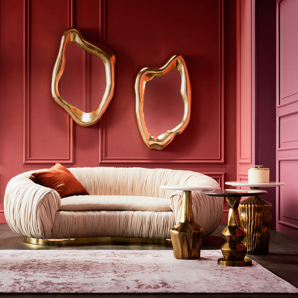 Cremefarbenes Sofa mit goldenem Sockel, zwei abstrakte Spiegel in Gold, Beistelltische und rot-rosa Wände in einem eleganten Raum.