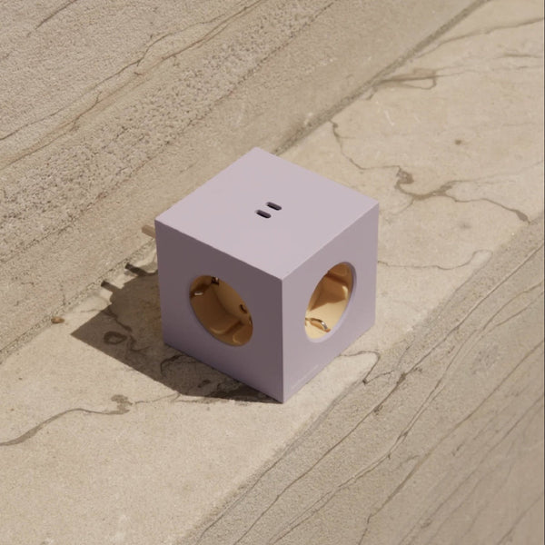 Eine lavendelfarbene, würfelförmige Steckdose mit drei runden Steckdosen liegt auf einer beigen Steinfläche im Sonnenlicht.