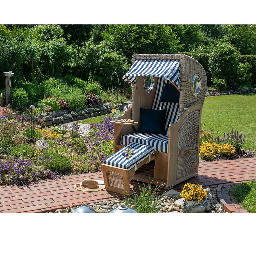 Ein Strandkorb mit blau-weiß gestreiften Kissen und Baldachin steht auf einem gemauerten Weg in einem Garten, umgeben von Blumen und Grünzeug.