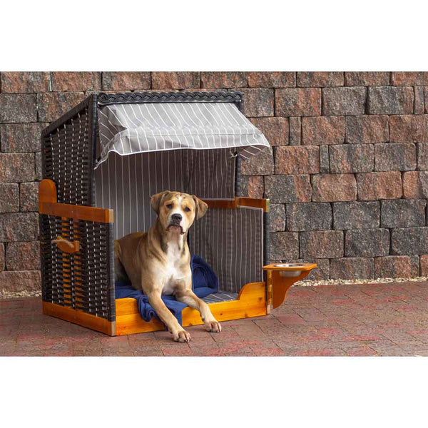 Ein brauner Hund sitzt in einer schattigen Hundehütte aus Holz mit einem gestreiften Vordach, einer blauen Decke im Inneren und einer Backsteinmauer im Hintergrund.
