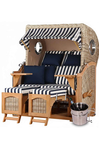 Strandkorb mit blau-weiß gestreiften Kissen, zwei ausklappbaren Fußstützen und einem Metall-Eiskübel mit einer Flasche daneben.