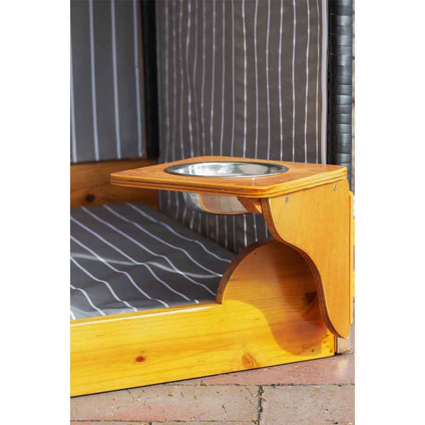 Ein hölzernes Bettgestell für Haustiere mit einem eingebauten erhöhten Regal, das einen Futter- oder Wassernapf aus Metall enthält und auf einer gefliesten Oberfläche steht.