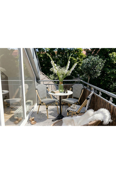 Ein kleiner Balkon mit zwei gemusterten Stühlen, einem runden Tisch mit einer Vase mit weißen Blumen, einer Topfpflanze und einem Blick auf die Bäume im Hintergrund.