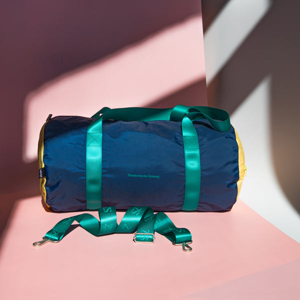 Tasche Weekender blau und  an den beiden Seiten gelb mit grünen Henkeln und Logo Süddeutsche Zeitung auf rosa Hintergrund 