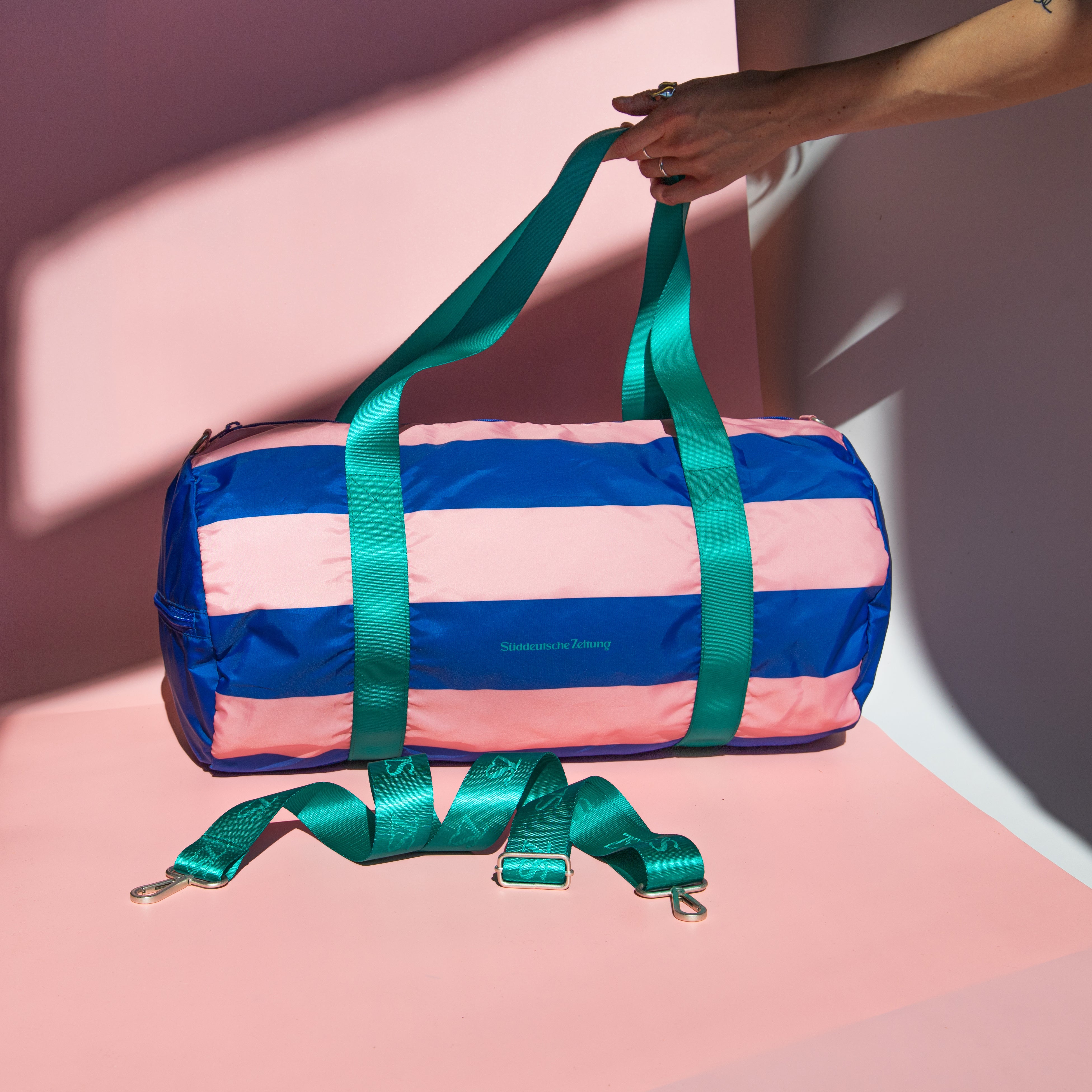 Tasche Weekender rosa blau gestreift  mit grünen Henkeln und Logo Süddeutsche Zeitung auf rosa Hintergrund. Eine Hand hält die Henkel der Tasche 