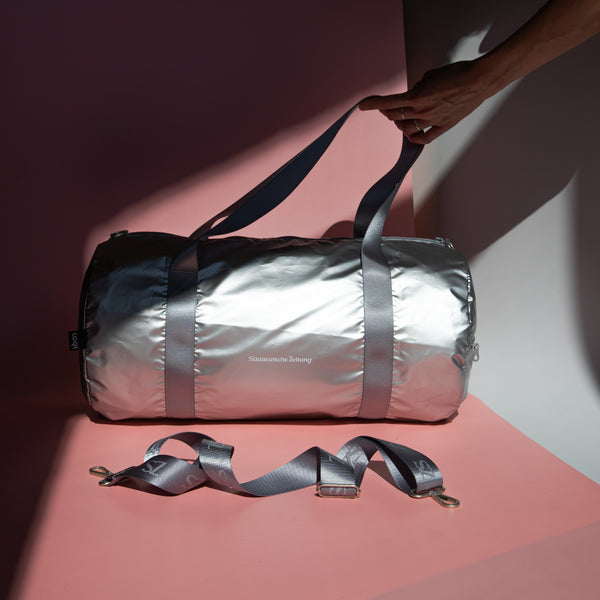 Tasche Weekender silber mit silbernen Henkel und Logo Süddeutsche Zeitung auf rosa Hintergrund ein Hand im Bild hält die Henkel der Tasche 