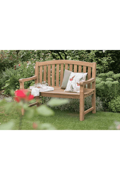 Hölzerne Gartenbank mit zwei gemusterten Kissen, einem Glaskrug, zwei Gläsern und einem gefalteten weißen Tuch, auf Gras mit grünen Pflanzen und Blumen im Hintergrund.