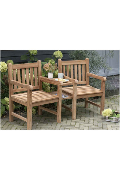 Zwei hölzerne Gartenstühle, die durch einen kleinen Tisch verbunden sind, stehen auf einer gepflasterten Terrasse mit Pflanzen und Blumen im Hintergrund.